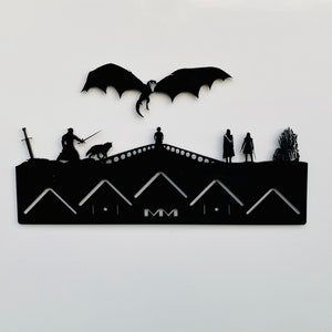 May include: Black metal wall decor featuring a silhouette of a dragon flying above a castle with characters from the popular TV show Game of Thrones. The text "MM" is visible on the castle wall.