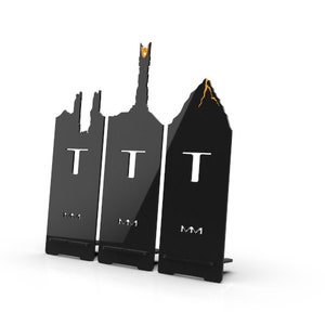 May include: Black metal silhouette wall decor featuring the iconic towers of Minas Tirith, the tower of Barad-dûr, and Mount Doom from the Lord of the Rings. The silhouette is made of three separate pieces, each with the letter "T" cut out.