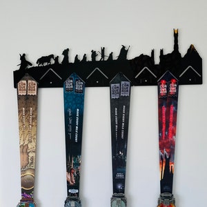 Puede incluir: Un soporte de pared de metal negro con forma de silueta de los personajes de El Señor de los Anillos y el Monte del Destino. Cuatro medallas de carrera coloridas cuelgan de los ganchos. Las medallas presentan imágenes de las películas de El Señor de los Anillos y el texto "CONQUER ROR" y "MAKE EVERY MILE COUNT".