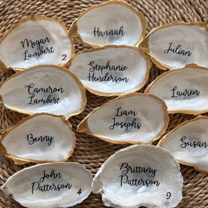 May include: A collection of oyster shells with gold-painted edges, each inscribed with a name in black script. The shells are arranged on a woven, natural-toned surface, ideal for place settings or decorative use.
