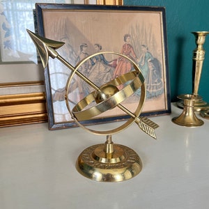 May include: A brass armillary sphere with an arrow through a ring, mounted on a circular base. The base has engraved markings. The sphere is displayed on a white surface, with framed artwork in the background.