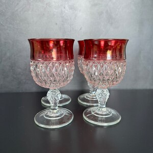 May include: Four vintage wine glasses with ruby red rims and diamond-cut patterned bowls. The clear glass stems have a decorative design and sit on round bases. The glasses are arranged on a dark surface against a gray background.