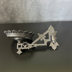 May include: An antique silver-toned wall sconce with an ornate design. The sconce features a decorative bowl-shaped holder and an elaborate bracket with intricate detailing. The sconce is designed to be mounted on a wall and has a vintage aesthetic.