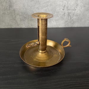 May include: A vintage brass candlestick holder with a round base, a central column, and a small handle. The candlestick holder has a classic design and is made of a shiny, golden-colored metal. The handle is small and curved.