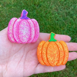 May include: Two pumpkin-shaped ornaments with a glittery finish. One pumpkin is bright pink with a purple stem, and the other is vibrant orange with a green stem. Both items feature vertical ridges and a textured, sparkling surface throughout.