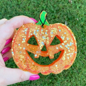 May include: A bright orange jack-o'-lantern ornament crafted from resin with a textured surface embedded with large, reflective gold and silver glitter. The pumpkin features a carved face with triangular eyes and a jagged mouth, topped with a green stem.