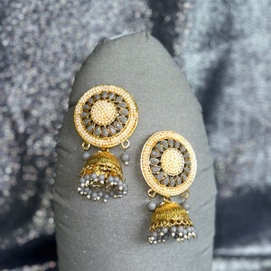 May include: A pair of gold earrings with gray and white beads. The earrings have a round design with a floral pattern and a dangling bell-shaped charm.