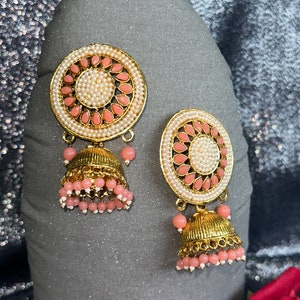 May include: A pair of gold earrings with pink and white beads. The earrings have a round, decorative top with a floral design and a dangling bell-shaped charm.