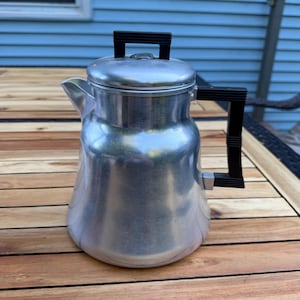 May include: A silver-coloured aluminium percolator coffee pot with a wide base and a tapered body. It features a black plastic handle on the side and a matching black plastic knob on the removable lid. The spout is integrated into the upper body.