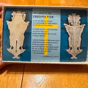 May include: A rectangular box containing two off-white, symmetrical fish bone structures resembling a crucifix. Between them is a blue card with the text The Legend of the CRUCIFIX FISH and a yellow cross graphic, detailing the story by Conrad S. Lantz.