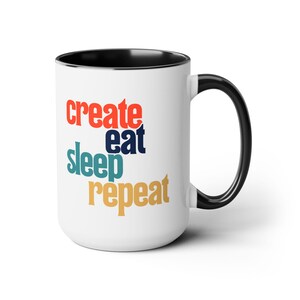 May include: White ceramic coffee mug with a black rim and handle. The mug features the text "create eat sleep repeat" in colorful, stylized lettering.