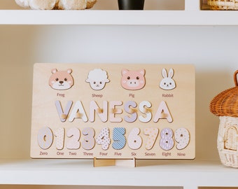 May include: A wooden educational toy with a pink, purple, and blue color scheme. The toy features a name puzzle with the name "VANESSA" and a number puzzle with the numbers 0-9. The toy also includes animal illustrations of a frog, sheep, pig, and rabbit.