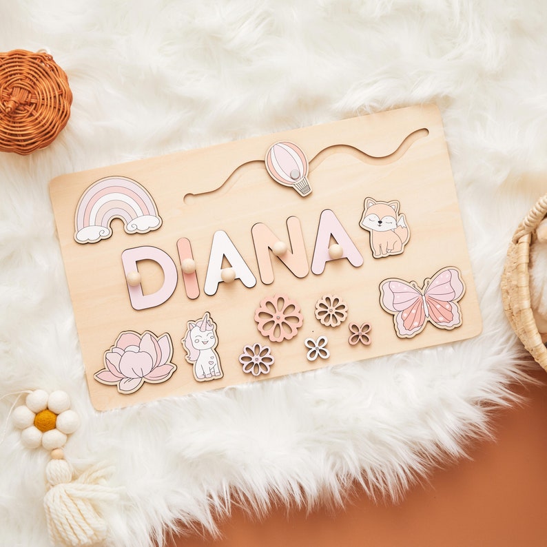 May include: Wooden name puzzle with the name "DIANA" in pink and white letters. The puzzle features a rainbow, hot air balloon, fox, butterfly, unicorn, and flower designs. The puzzle is on a light wood base.