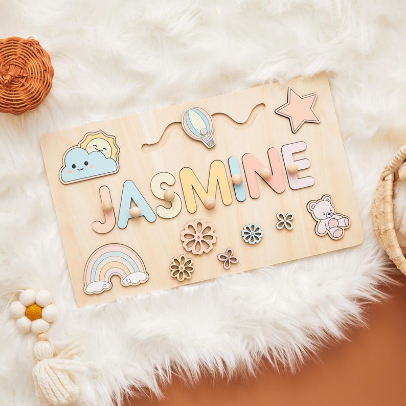 May include: Wooden name puzzle with the name "JASMINE" in pastel colors. The puzzle includes a rainbow, cloud, sun, hot air balloon, star, flowers, and a teddy bear. The puzzle is made of light-colored wood.