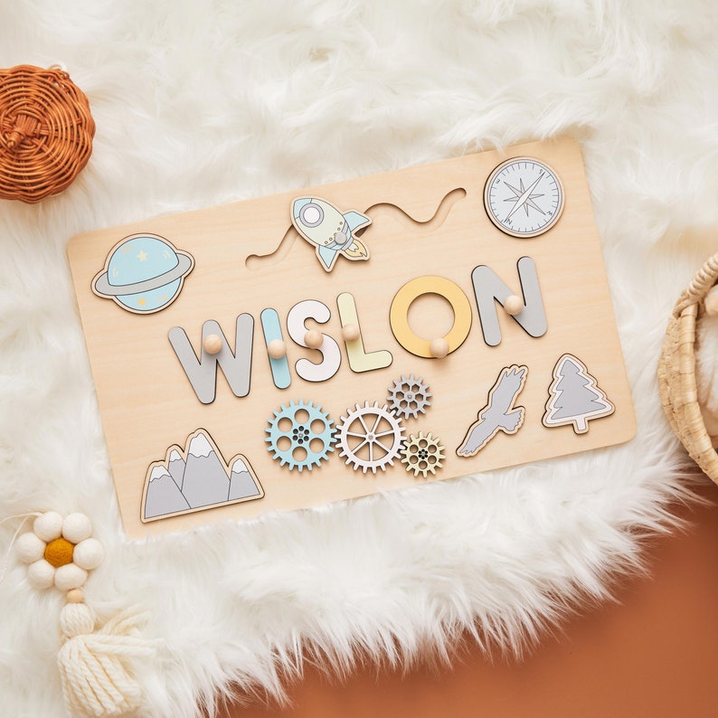 May include: A wooden name puzzle with the name "WISLON" in gray, yellow, and blue letters. The puzzle includes space-themed shapes like a rocket, planet, and compass, along with gears, mountains, and a tree. The puzzle is on a white fluffy surface.