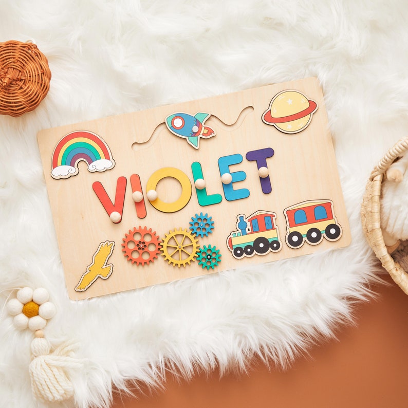 May include: Wooden name puzzle with the name "VIOLET" in colorful letters. The puzzle includes a rainbow, rocket, planet, gears, train, and a bird. The puzzle is on a light wood base.