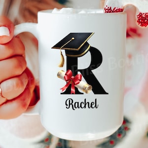 Puede incluir: Taza de cerámica blanca con la letra negra "R", un gorro de graduación y un diseño de diploma. El nombre "Rachel" está impreso debajo. Una galleta roja y blanca está colocada en el borde.
