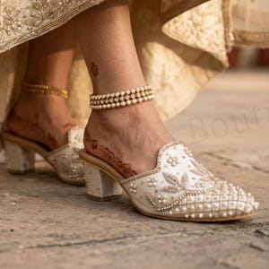 May include: Ivory-colored, embellished mules with a low block heel. The shoes feature intricate beadwork and pearl accents. A gold anklet and a pearl anklet are visible. The image shows a close-up of the shoes and ankles.