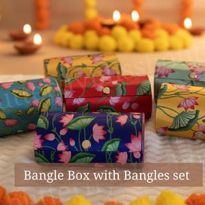 May include: A collection of colorful bangle boxes with floral patterns. The boxes are in various colors, including blue, red, and yellow, and are adorned with pink lotus flowers and green leaves. The text "Bangle Box with Bangles set" is visible.