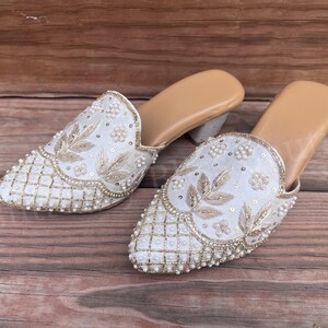 May include: A pair of white embroidered mules with a pointed toe. The shoes feature intricate gold thread and pearl embellishments. The heels are a light gray color, and the soles are tan.
