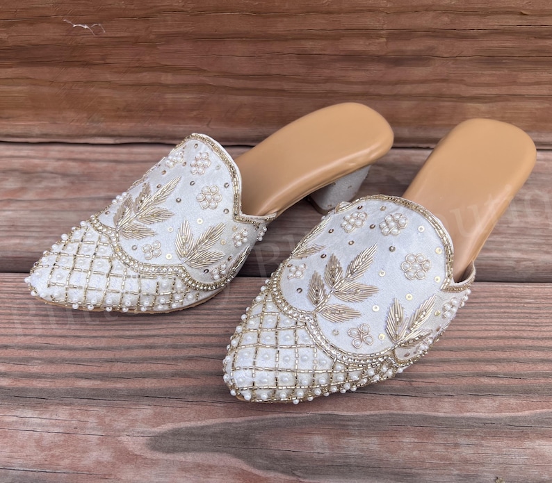 May include: A pair of white embroidered mules with a pointed toe. The shoes feature intricate gold thread and pearl embellishments. The heels are a light gray color, and the soles are tan.