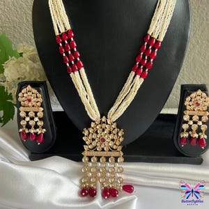 May include: A gold necklace with a large pendant featuring a red stone surrounded by white stones. The necklace is made of white beads and red beads. The pendant is attached to a chain of white beads. The necklace is accompanied by a pair of earrings with a similar design.
