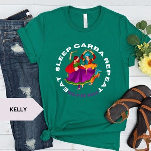 Puede incluir: Una camiseta verde con un gr&aacute;fico circular blanco que representa a dos personas bailando con palos. El texto alrededor del c&iacute;rculo dice "Eat Sleep Garba Repeat Halo Re Halo".
