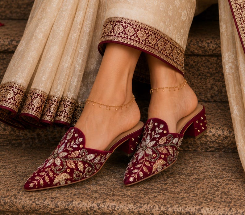 May include: Burgundy velvet mules with intricate floral embroidery and gold accents. The shoes have a pointed toe and a block heel. A gold anklet with small beads is worn on each ankle. The background includes a cream-colored fabric with gold and burgundy trim.