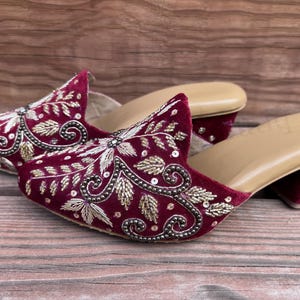 May include: A pair of burgundy velvet mules with intricate gold and silver embroidery. The open-toe shoes have a small heel and a tan insole. The detailed floral design is embellished with sequins and beads.
