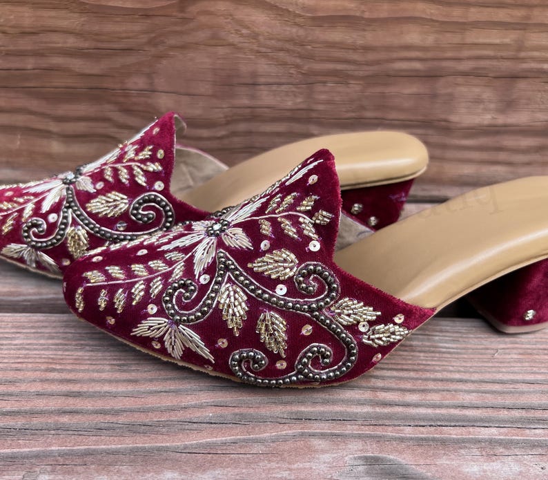 May include: A pair of burgundy velvet mules with intricate gold and silver embroidery. The open-toe shoes have a small heel and a tan insole. The detailed floral design is embellished with sequins and beads.