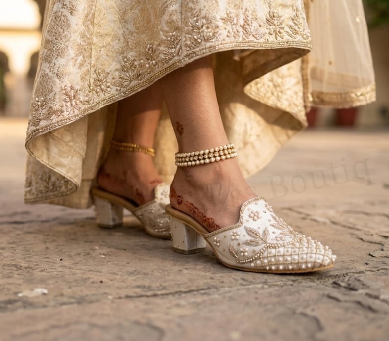 May include: Ivory-colored, embellished mules with a low block heel. The shoes feature intricate beadwork and pearl accents. A gold anklet and a pearl anklet are visible. The image shows a close-up of the shoes and ankles.
