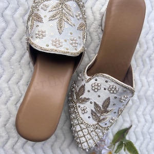 May include: A pair of white embroidered slip-on shoes. The shoes feature intricate gold leaf and floral designs, with pearl and sequin embellishments. The soles are a light brown color.