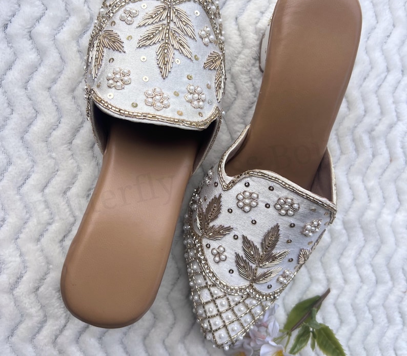 May include: A pair of white embroidered slip-on shoes. The shoes feature intricate gold leaf and floral designs, with pearl and sequin embellishments. The soles are a light brown color.