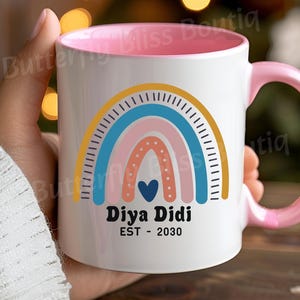 Könnte beinhalten: Weißer Keramikbecher mit rosa Innenraum und Griff. Der Becher zeigt ein farbenfrohes Regenbogen-Design mit dem Text "Diya Didi EST - 2030" in Schwarz. Der Regenbogen umfasst gelbe, blaue und rosa Bögen mit einem blauen Herzen.
