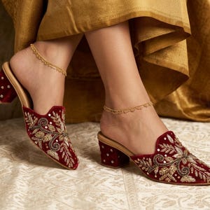 May include: Burgundy velvet mules with intricate gold and silver embroidery and sequin accents. The open-back shoes have a block heel and a gold-colored insole. A delicate gold anklet is worn on each ankle. The shoes are on a patterned surface.