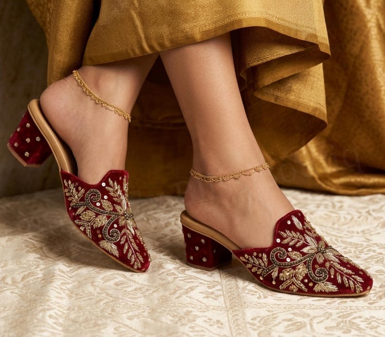 May include: Burgundy velvet mules with intricate gold and silver embroidery and sequin accents. The open-back shoes have a block heel and a gold-colored insole. A delicate gold anklet is worn on each ankle. The shoes are on a patterned surface.