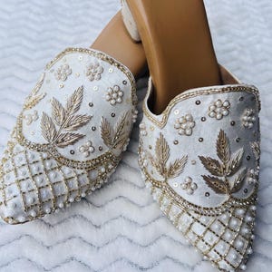 May include: A pair of white, ornate slip-on shoes. The shoes are heavily embellished with gold embroidery, sequins, and small pearls. The design includes floral and leaf motifs, with a grid pattern on the toe.