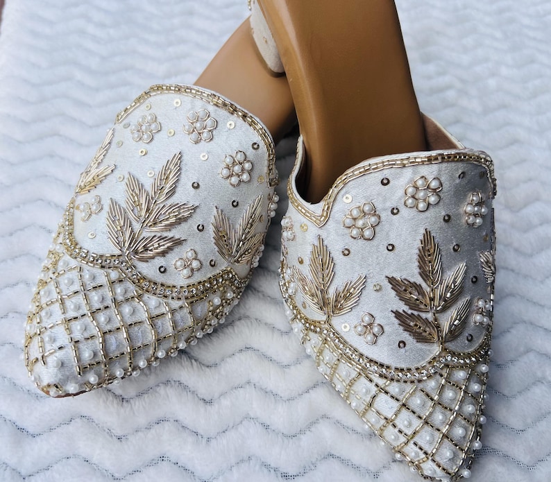 May include: A pair of white, ornate slip-on shoes. The shoes are heavily embellished with gold embroidery, sequins, and small pearls. The design includes floral and leaf motifs, with a grid pattern on the toe.