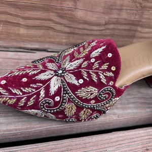 May include: Close-up of a burgundy velvet mule with intricate gold and silver embroidery. The shoe features a floral design with sequin accents and a small block heel. The sole is a light tan color.