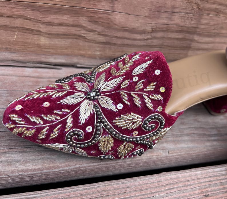 May include: Close-up of a burgundy velvet mule with intricate gold and silver embroidery. The shoe features a floral design with sequin accents and a small block heel. The sole is a light tan color.