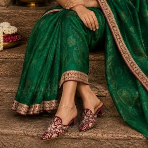 May include: A person wearing a green sari with gold trim and embroidered maroon mules. The sari has a gold paisley pattern. The mules have a pointed toe and a small heel. A gold anklet is visible.