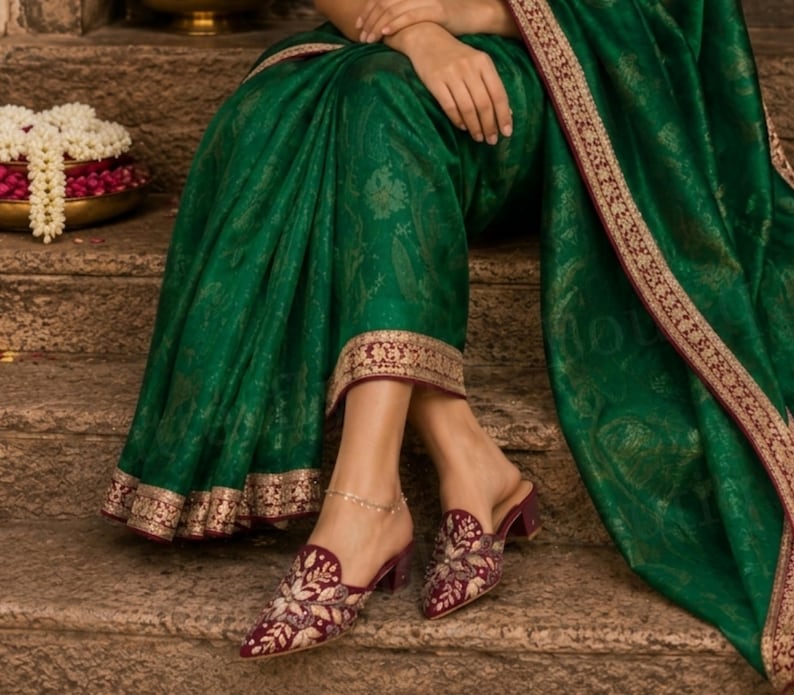 May include: A person wearing a green sari with gold trim and embroidered maroon mules. The sari has a gold paisley pattern. The mules have a pointed toe and a small heel. A gold anklet is visible.