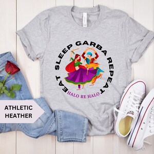 Puede incluir: Una camiseta gris con un gr&aacute;fico circular que muestra a dos personas bailando. El texto alrededor del c&iacute;rculo dice "Eat Sleep Garba Repeat" y "Halo Re Halo".