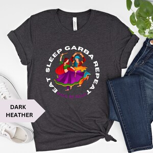 Puede incluir: Camiseta gris oscuro jaspeada con un gr&aacute;fico circular blanco que representa a una pareja bailando con palos. El texto alrededor del gr&aacute;fico dice "Eat Sleep Garba Repeat" con las palabras "Halo Re Halo" en la parte inferior.