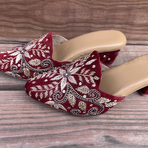 May include: A pair of burgundy velvet mules with intricate gold and silver embroidery. The open-toe shoes have a low heel and a tan insole, showcasing a floral design.