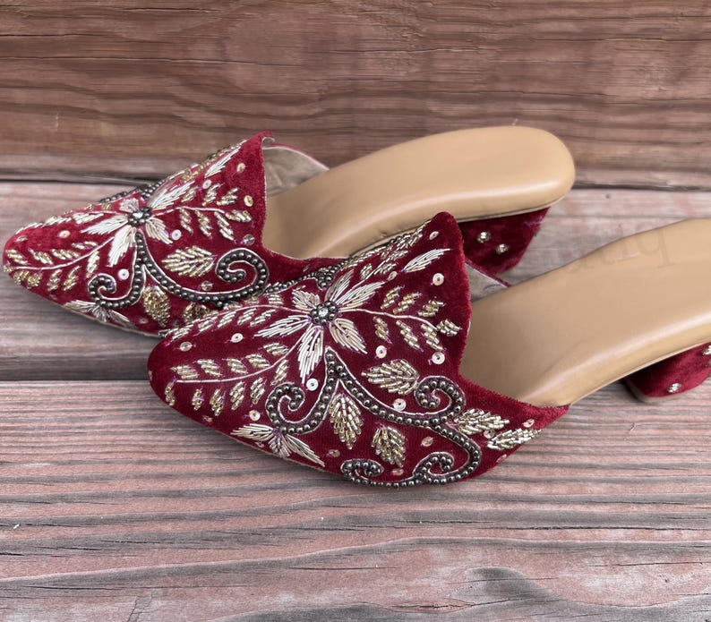May include: A pair of burgundy velvet mules with intricate gold and silver embroidery. The open-toe shoes have a low heel and a tan insole, showcasing a floral design.