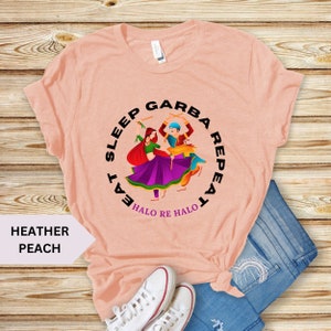 Puede incluir: Una camiseta de color melocot&oacute;n con un gr&aacute;fico de dos personas bailando con palos. El gr&aacute;fico est&aacute; en un c&iacute;rculo con el texto "Eat Sleep Garba Repeat" y "Halo Re Halo" debajo.