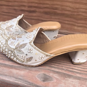 May include: A pair of ivory-colored, embellished mules with a low heel. The shoes feature intricate beadwork, floral embroidery, and gold accents. The interior is a light tan color, and the shoes are set on a wooden surface.