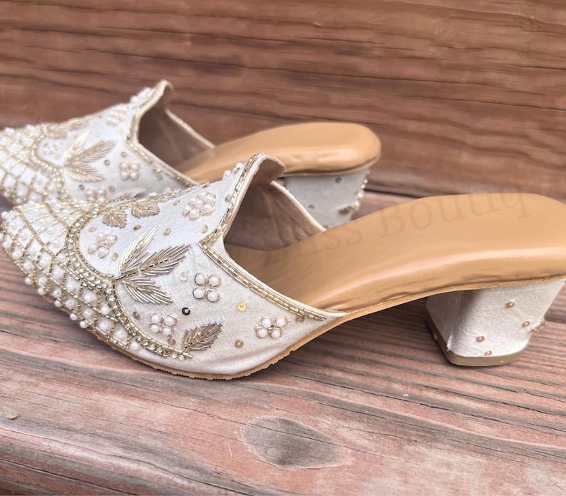 May include: A pair of ivory-colored, embellished mules with a low heel. The shoes feature intricate beadwork, floral embroidery, and gold accents. The interior is a light tan color, and the shoes are set on a wooden surface.