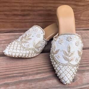 May include: A pair of white, slip-on mules with intricate embroidery and beadwork. The shoes feature gold-colored leaf and floral designs, with a grid pattern on the toe. The soles are a light tan color, and the shoes are set against a wooden background.
